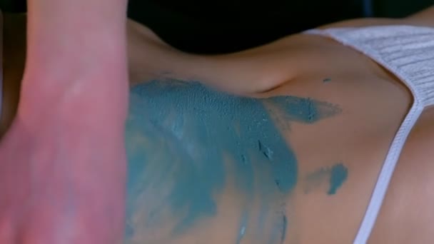 Cosmetologist homem aplicando barro azul no corpo das mulheres no abdômen, mãos de close-up. — Vídeo de Stock