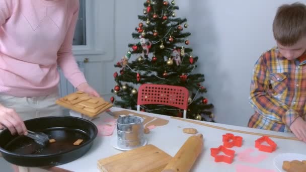 Mother and son puts parts of baked gingerbred house on wooden board using tongs. — Stock Video