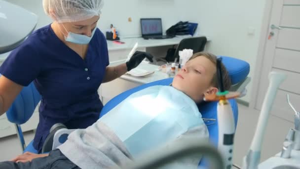 Barnpojken sitter i tandläkarstolen, doktorn förbereder sig för undersökning på kliniken. — Stockvideo