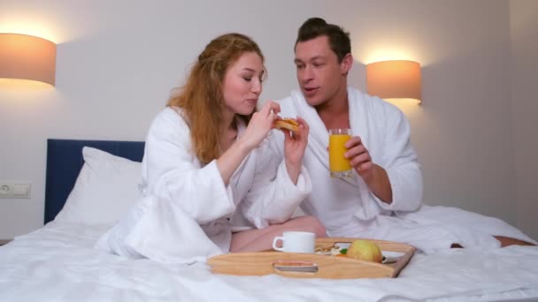 Mężczyzna i kobieta jedzą razem śniadanie siedząc w łóżku w pokoju hotelowym. — Wideo stockowe