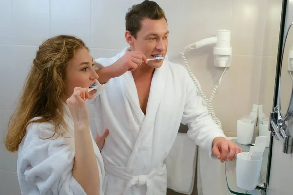 Homem e mulher em roupões de banho limpando os dentes juntos no banheiro antes do espelho. — Fotografia de Stock