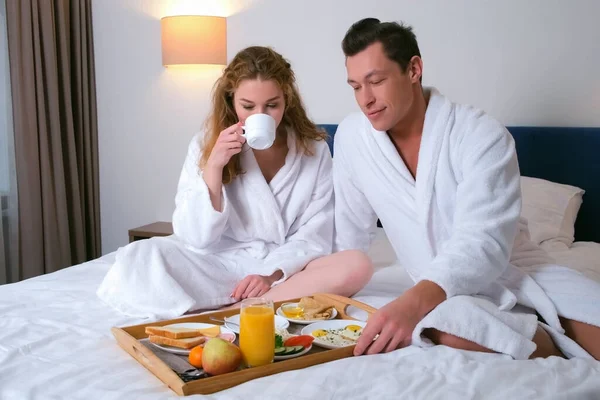 若いですカップルでバスローブで朝食を一緒にベッドでホテルの部屋. — ストック写真