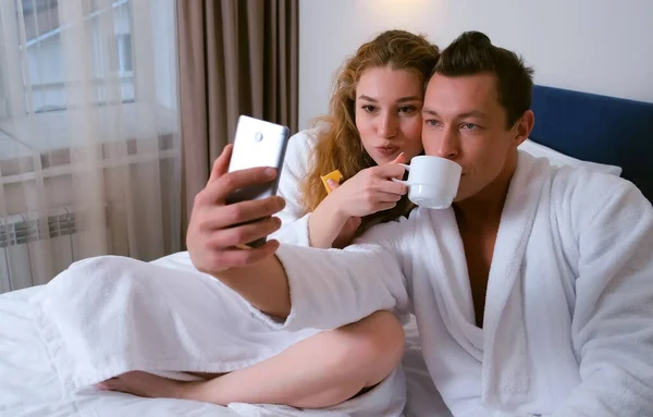 男と女でバスローブを着て自画撮り写真上のスマートフォンで食べる. — ストック写真