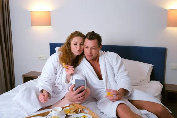 男性と女性でバスローブを着てホテルで食べるスマートフォンで写真を撮る. — ストック写真
