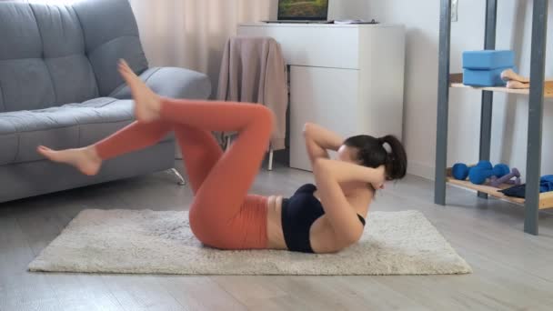 Jonge vrouw in sportkleding doet buikspieroefeningen buigende benen op tapijt thuis. — Stockvideo