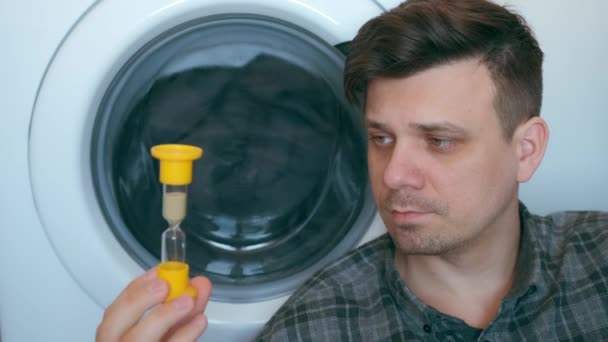 Man met zandglas wacht op de wasmachine met grijze sprei erin. — Stockvideo