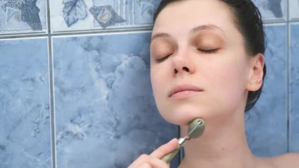 Mujer joven es relajante tumbado en el baño y haciendo masaje facial con rodillo de jade. — Vídeo de stock
