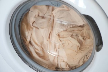 İçinde bej çarşaf olan çamaşır makinesi çalışıyor, evde çamaşır yıkılıyor..