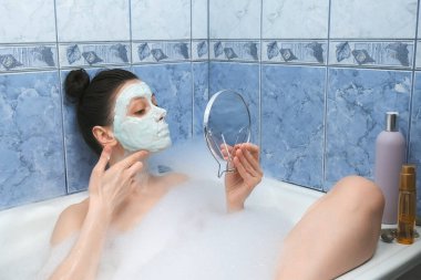Kadın küvete köpüklü aljinat maskesi takıyor. Küçük aynaya bakıyor..