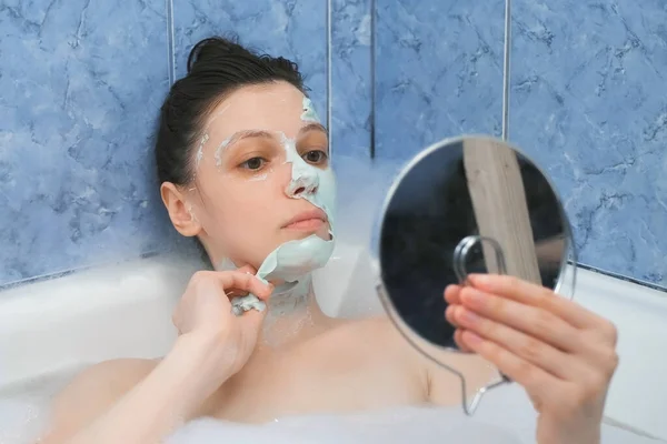 Young woman is removing alginate mask from her face lying in bathroom with foam. — Stock Photo, Image