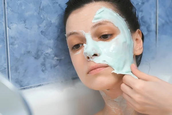 Mujer joven está eliminando la máscara de alginato de su cara acostada en el baño con espuma. — Foto de Stock