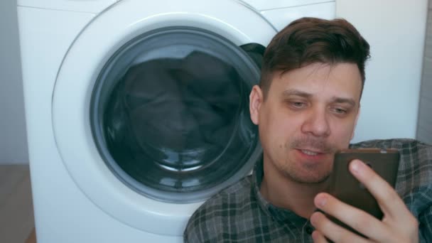 Mann wartet mit Tagesdecke auf Waschmaschine und spricht auf Smartphone. — Stockvideo