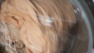 İçinde bej çarşaf olan çamaşır makinesi çalışıyor, evde çamaşır yıkılıyor..