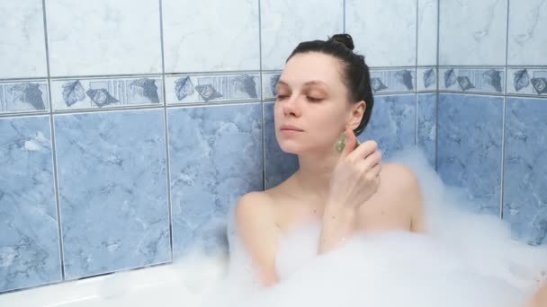 Young woman is relaxing lying in bath doing face massage using gouache scraper. — Stock Video
