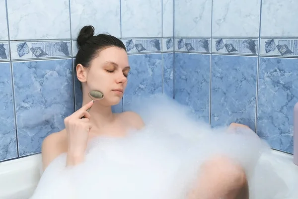 Young woman is relaxing lying in bath and doing face massage using jade roller. — Stock Photo, Image