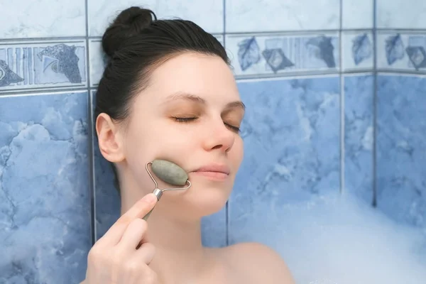 Mujer joven es relajante tumbado en el baño y haciendo masaje facial con rodillo de jade. — Foto de Stock