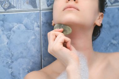 Genç kadın banyoda uzanıp, guaj kazıyıcı kullanarak boyun masajı yapıyor..