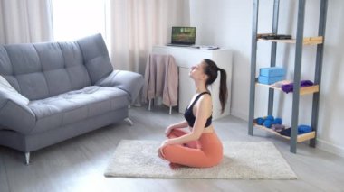 Yogada arka bükme egzersizi yapan genç bir kadın. Evde minderde oturuyor, yan gözle bakıyor..
