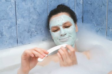 Genç bir kadın banyoda yüzünde aljinat maskesiyle tırnaklarını törpülüyor..