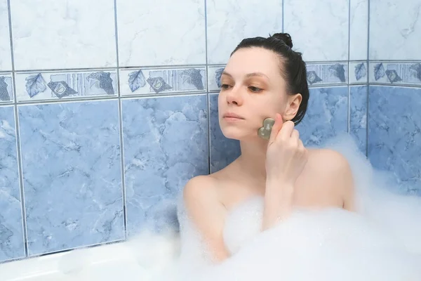 Mujer joven es relajante tumbado en el baño haciendo masaje facial utilizando gouache rascador. — Foto de Stock