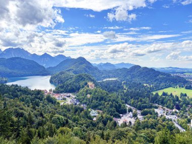 Neuschwanstein Kalesi 'nin yukarısından, Alpsee Gölü yakınlarındaki Bavyera Alpleri' nin dağ ormanlarıyla çevrili bir tepenin tepesinde, mavi, parçalı bulutlu bir gökyüzüne karşı gizlenmiş bir panorama..