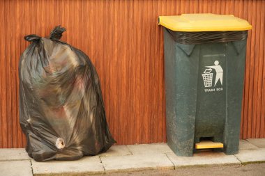Büyük siyah bir çöp torbası yeşil bir çöp kutusunun yanında oturur. Kaldırımda sarı bir kapağı olan, kahverengi ahşap bir arka planda kurulmuş..