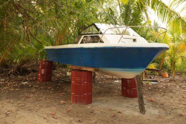 Mavi ve beyaz bir tekne tropikal bir ortamda kırmızı fıçılarda oturur, palmiye ağaçları ve yeşilliklerle çevrili, terk edilme ve ihmal hissi verir..