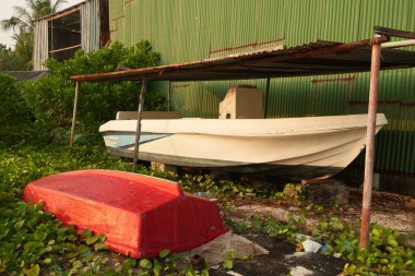 Bereketli bitki örtüsü ve ön planda kırmızı tekne gövdesi ile çevrili yeşil kıvrımlı metal bir yapının yakınındaki bir barınağın altında teknelerin olduğu sakin ama ihmal edilmiş bir sahne..