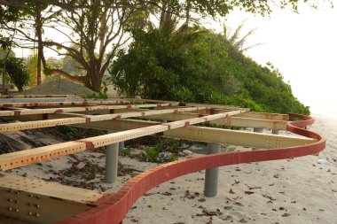 Ağaçlar ve yeşilliklerle çevrili, kıvrımlı kırmızı tuğlalı bir kenarı ve metal kirişleri olan kumlu bir sahilde ahşap bir yapı yapım aşamasındadır ve arka planda parlak bir gökyüzü vardır..