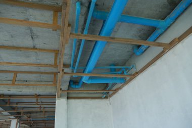 Bir inşaat sahasının içinde tavanda mavi borular ve ahşap kirişler var. Kısmen bitmiş beyaz bir duvarın yanında binanın altyapısını gösteriyor..