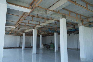 Parlak zemini, beyaz duvarları ve tavanda açık ahşap kirişleri ve boruları olan geniş, boş bir oda. Arka planda birkaç beyaz sütun ve inşaat malzemesi var..