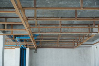 Ahşap kirişler ve metal parantezlerden oluşan ve içinden mavi boruların geçtiği beton duvarlar ve tavanlardan oluşan açık tavanlı bir oda..