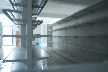 Beyaz duvarları ve zeminleri olan iyi ışıklandırılmış, boş bir perakende alanında, arka planda mavi bir kova ve karton kutularla stok ve organizasyon bekleyen, sıra sıra, metal raf üniteleri yer alıyor..