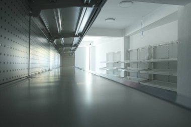 Sağında beyaz raflar ve gri bir zemini olan uzun, boş bir perakende mağaza koridoru, parlak ışıklandırması ve temiz, minimalist estetiği olan modern bir iç tasarımı içeriyor..