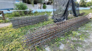 İnşaat alanında yeni bir inşaat projesinde kullanılmayı bekleyen çimenli bir zeminde duran güçlendirilmiş çelik çubuklar (inşaat çubuğu) ve prefabrik tel örgü yapıları yığını.