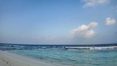 Okyanusun nazik, berrak turkuaz dalgaları yumuşak, kumlu bir kumsalla buluşur. Dağınık mavi bulutlarla dolu sakin bir gökyüzü altında tropikal bir manzara yaratır..