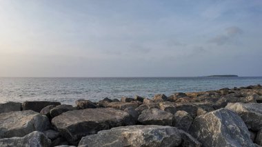 Ufukta soluk bir ada olan huzurlu bir okyanus manzarası ön planda büyük ve yıpranmış kayalar topluluğu tarafından çerçevelenmiş..
