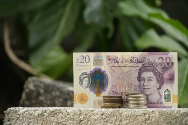 Bir İngiliz 20 poundluk banknot ve bozuk para yığınları beton bir blok üzerine yerleştirilmiş, solunda bulanık yeşil bir arka plan ve başka bir somut element var..