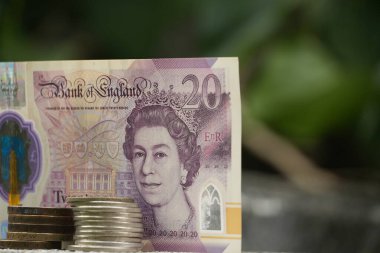 Bozuk para yığınlarıyla dolu 20 poundluk bir İngiliz banknotu beton bir yüzey üzerinde durmakta. Güzelce çerçevelenmiş yeşil bir arka plan, doğadaki mali istikrarın bir göstergesi olarak..