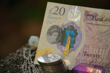 İngiliz 20 pound 'luk banknot, dokulu taş bir yüzeye karşı dimdik duruyor ve ilgi çekici bir görüntü yaratıyor. Yanında parlak, yanardöner renkleri olan gümüş bir zincir takılı kristal bir kolye yatar.,