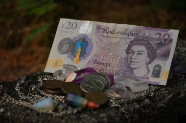 Bu resim, dağınık madeni paraların, çeşitli kristallerin ve arka planda yeşil bir ipucunun bulunduğu kırsal bir taşın üzerindeki gümüş bir zincirin arasında yirmi pound değerinde bir banknot sunuyor..