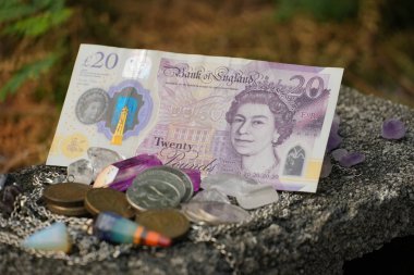 İngiliz sikkelerinin arasına dikkatlice yerleştirilmiş 20 poundluk bir banknot ve desenli bir taş yüzeyde değerli ve doğal elementleri yansıtan temiz ve mor kristallerin ilginç bir karışımı..