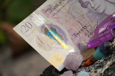 20 poundluk banknotun sol kısmını vurgulayan yakın plan, köşegen bir çekim, canlı renkli bir cazibe, madeni paralar ve desenli taş bir yüzeye dağılmış berrak kristaller..