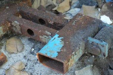 Üzerinde mavi boya kalıntıları olan ağır, paslı metal kirişler kumlu zemine atılmış, kuru yapraklarla çevrili, zamanla çürüyen ve endüstriyel ihmalkarlık gösteren.