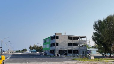 Bu görüntü, inşaat halindeki çok katlı beton bir binayı, parlak mavi gökyüzünün altında palmiye ağaçları bulunan geniş ve temiz bir yolun yanında gösteriyor..