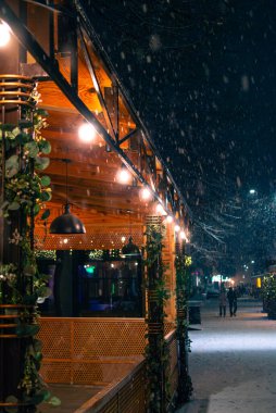 Şenlikli şehir ışığında süslü bir kafe terasının yanında gece kar yağışı