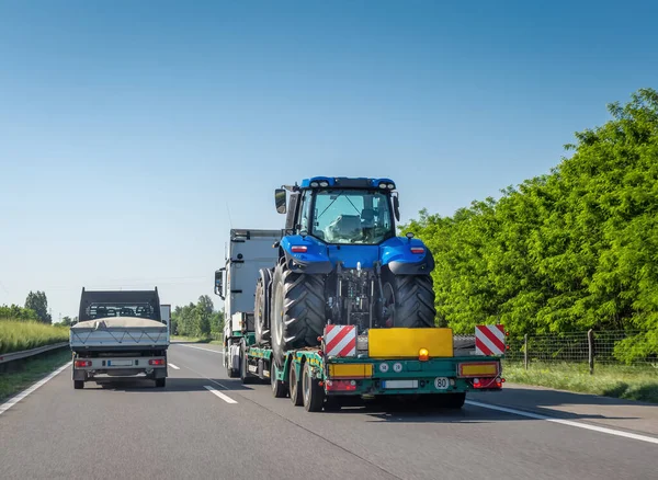 Karayolu üzerinde ağır makineleri taşımak için uzun kamyon platformunda yeni mavi traktör. Tarım makinelerinin nakliyesi