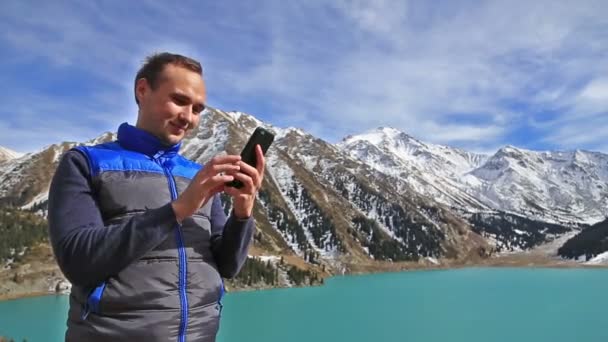 肯定的な笑顔の男の肖像は美しい山の湖の風景の背後にある使用スマートフォンを使用します — ストック動画