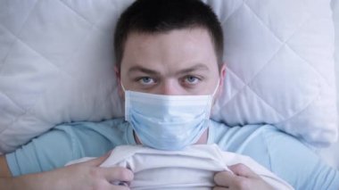 Tıbbi maske takan hasta bir adam koruyucu maske takarak yatakta yatıyor. Ateş, baş ağrısı ve bulantı belirtilerdir. Coronavirus Covid-19. Sağlık kavramı