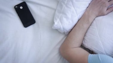 Genç adam uyandı, telefona baktı ve tekrar uyumaya başladı. Telefondaki çalar saat. Yorgun bir adam yatak odasında yatakta uyanıyor. Modern teknoloji kavramı